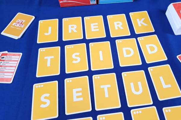 Wort-Ratespiel mit gelben Buchstabenkarten auf blauem Tisch bei der Queen-Games-Herbstneuheiten-Präsentation, im Hintergrund eine rote Queen-Games-Spielschachtel